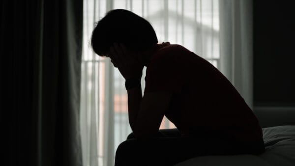 Silhouette of a person sitting on a bed with their head in their hands, appearing distressed in front of a window with curtains.