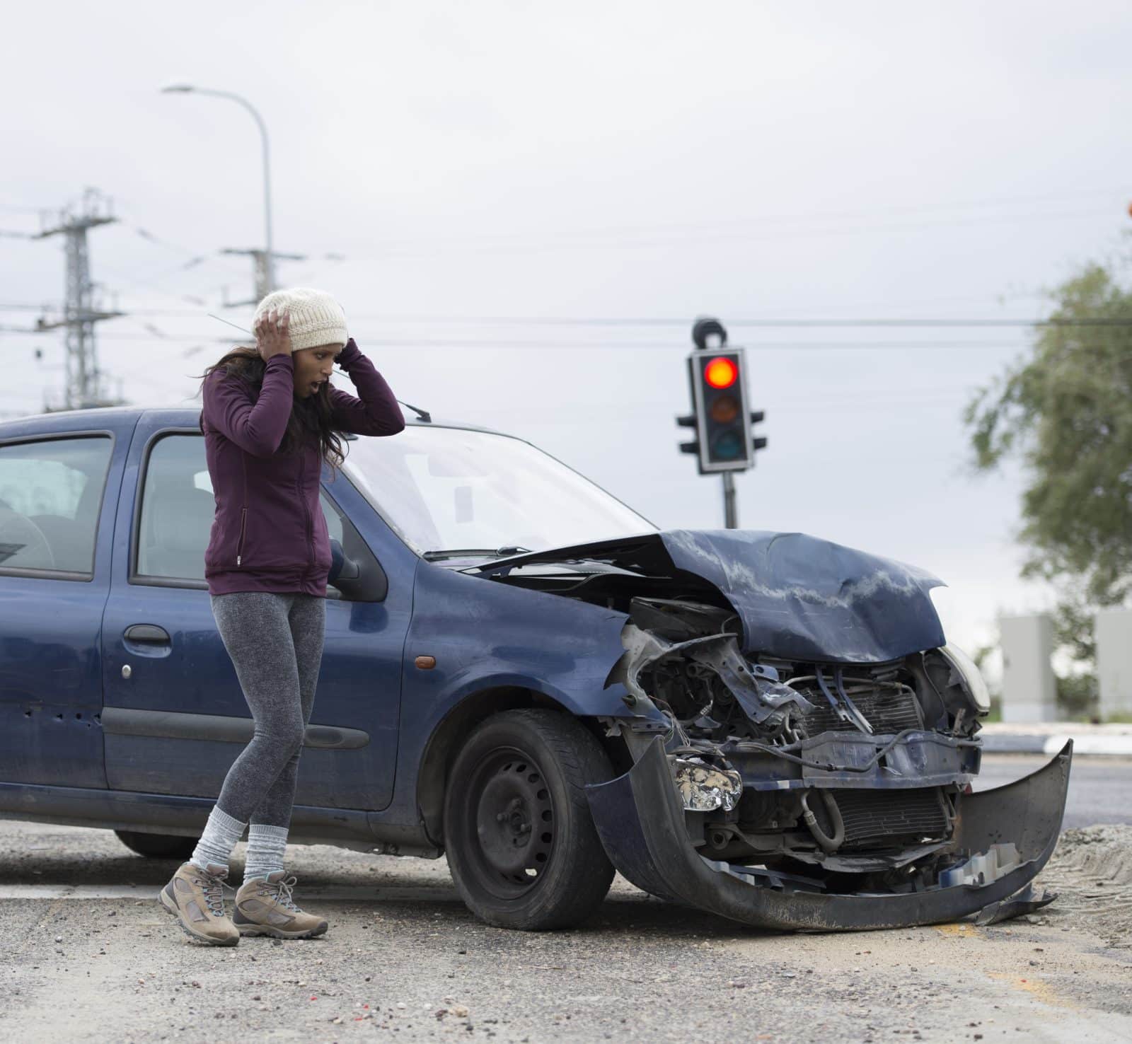 Woman with shocked face looking on her damaged car, holding head in hands. Car accident with frontal impact. Red traffic light is on background.