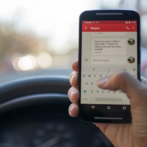 A driver holding a smartphone and typing a text message while driving, with one hand on the steering wheel.