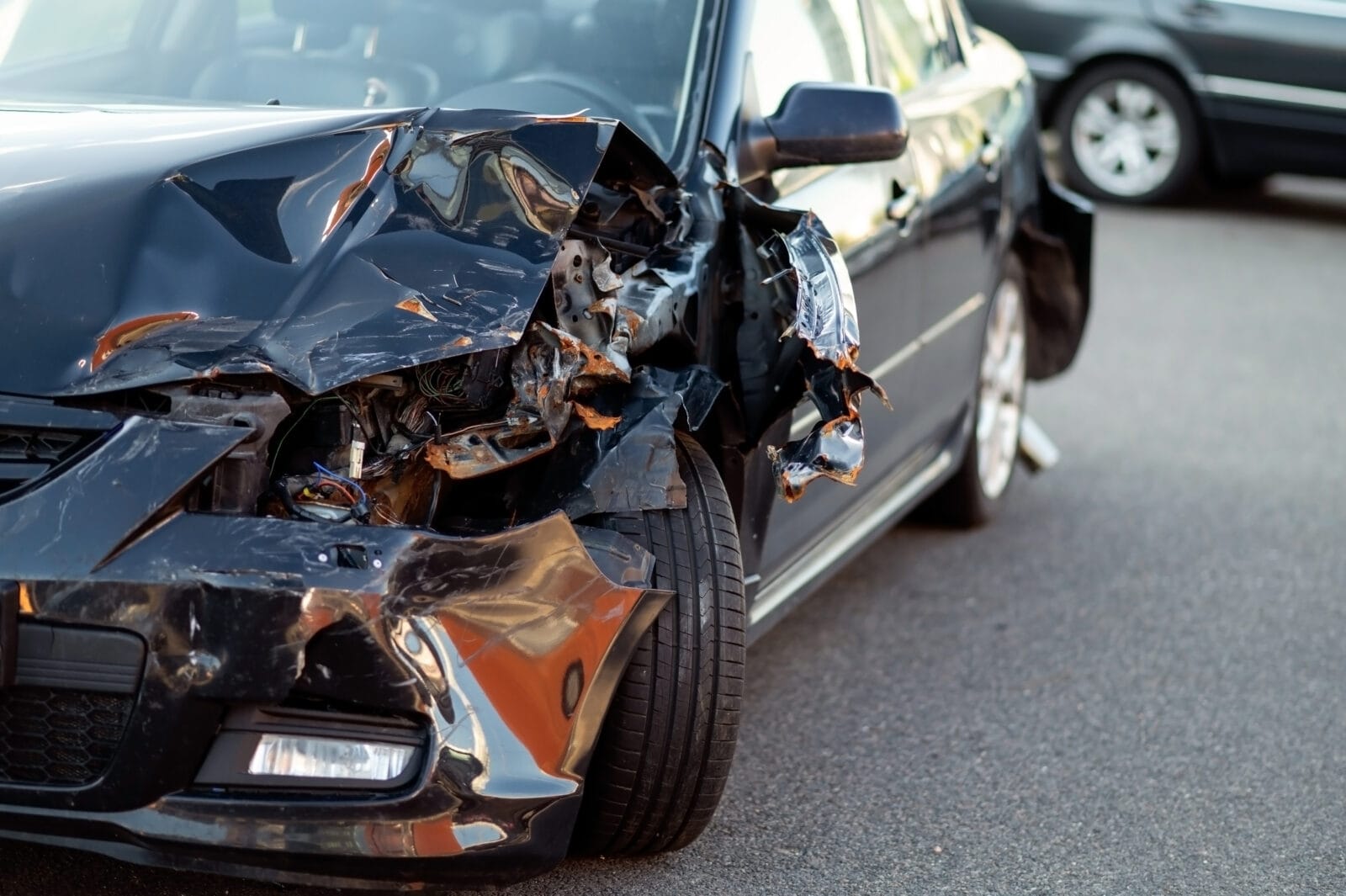 A black car with severe front-end damage sits on the road after a collision.