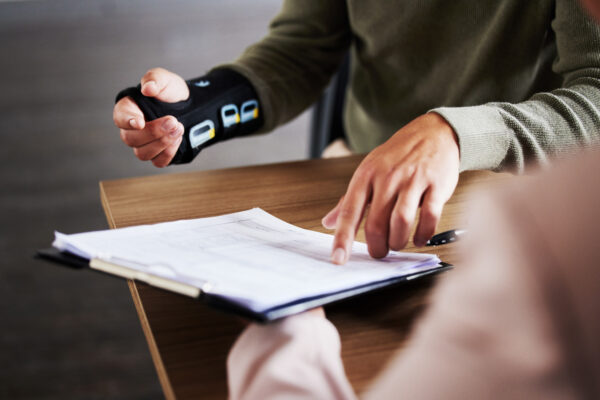 A person wearing a wrist brace sits at a desk and points to documents on a clipboard held up by someone else.