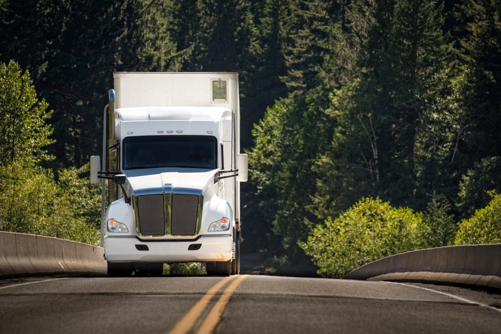 White semi-truck driving on a rural road surrounded by trees.
