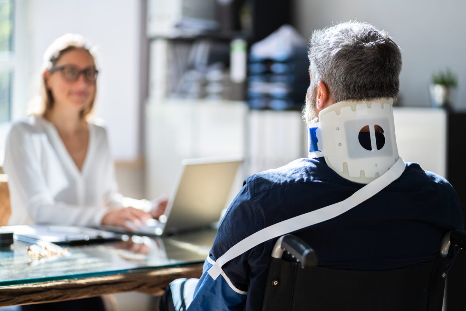 A man wearing a neck brace and seated in a wheelchair meets with a professional at a desk.