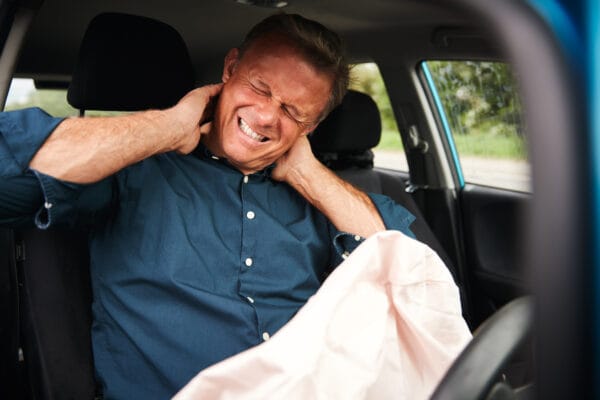 Man sitting in a car grimacing in pain while holding his neck after a car accident, with a deployed airbag visible.