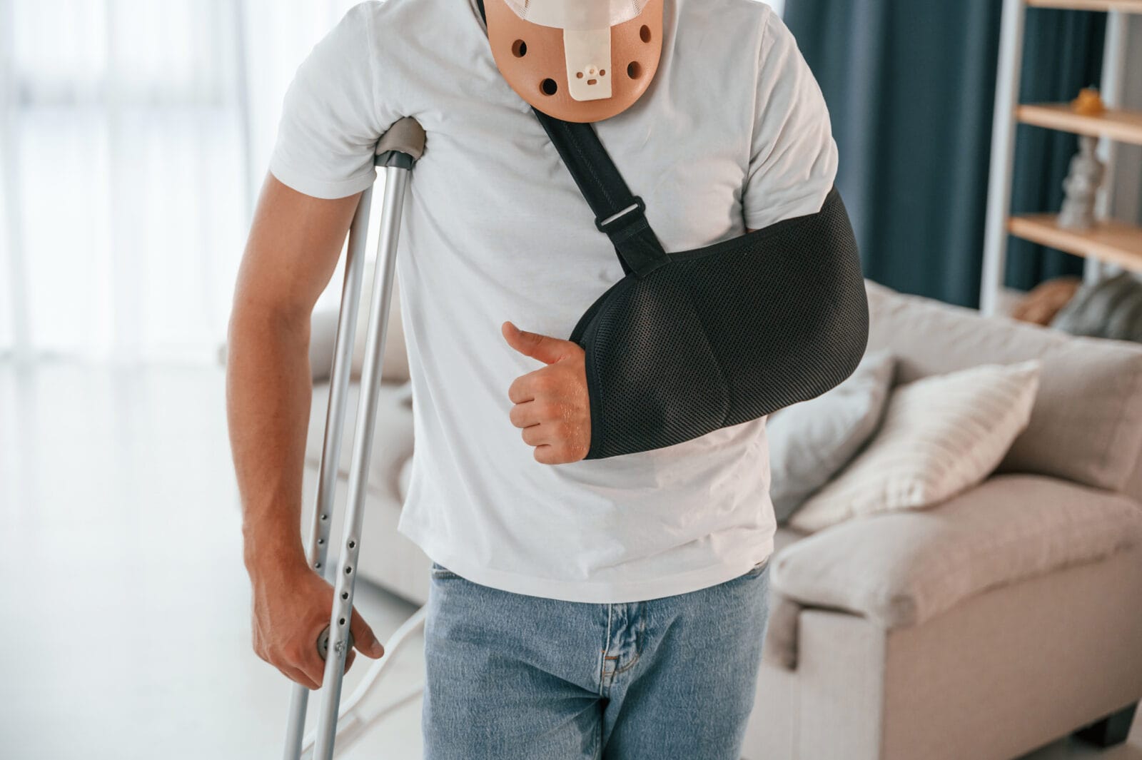 Man wearing a neck brace and arm sling using crutches while giving a thumbs-up gesture at home.